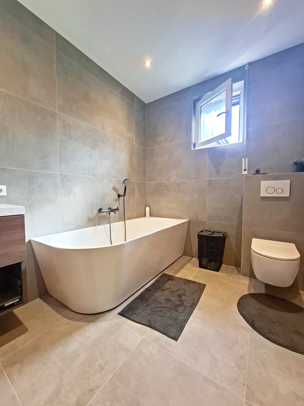 Moderne badkamer met vrijstaand ligbad, wandtoilet, grijze wandtegels, lichte vloertegels en raam.