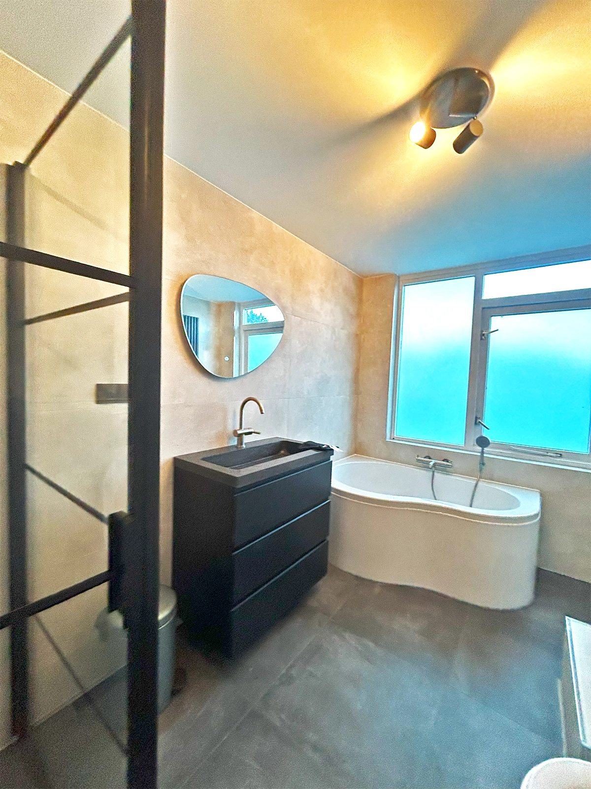 Moderne badkamer met ligbad, zwarte inloopdouche, zwart badkamermeubel, ronde spiegel en grote ramen.