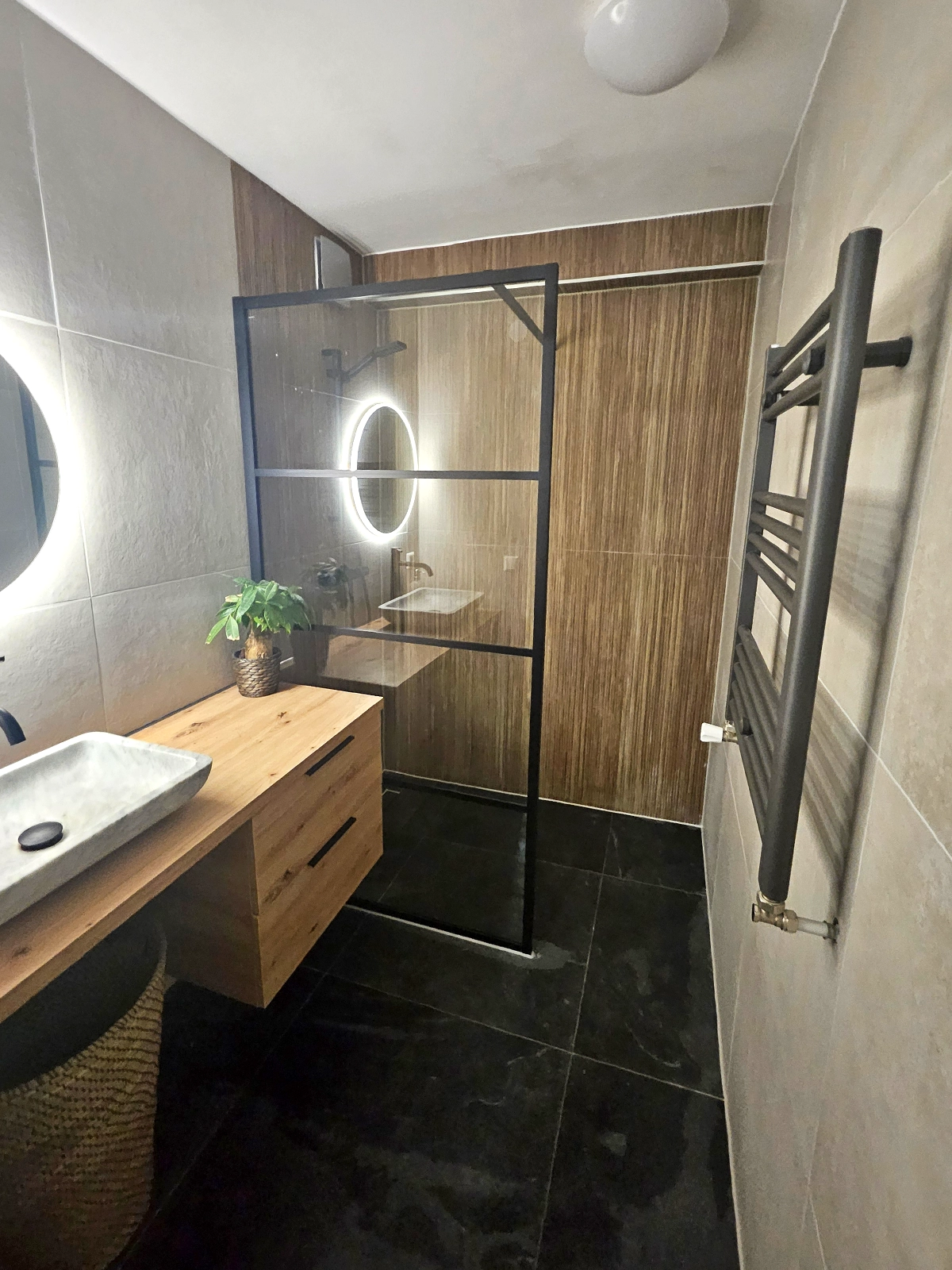 Moderne badkamer met inloopdouche, houtlook wandtegels, houten badkamermeubel, ronde verlichte spiegel en zwarte verwarming.