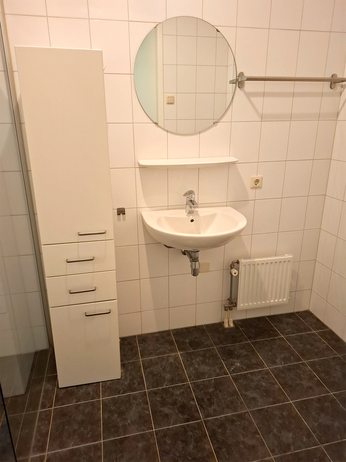 Badkamer met wastafel, ronde spiegel, hoge kast, radiator en witte wandtegels.