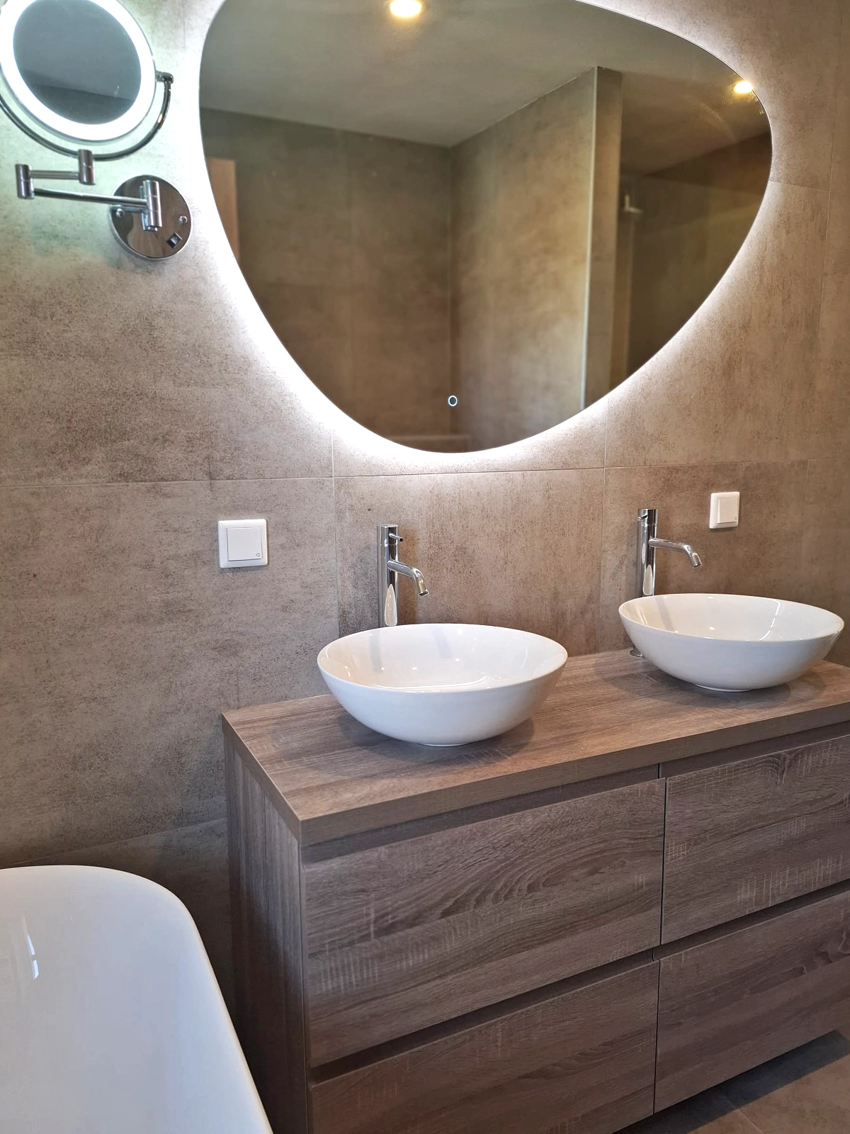 Moderne badkamer met dubbele waskommen, houten badkamermeubel, ovale verlichte spiegel, vrijstaand bad en grijze wandtegels.