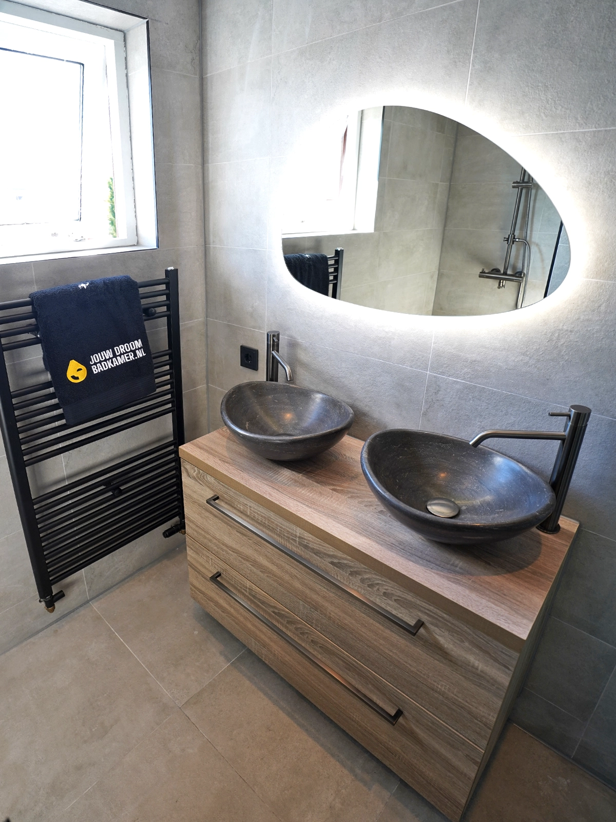 Moderne badkamer met dubbele waskommen, ovaal verlichte spiegel, houten badkamermeubel, zwarte kranen en zwarte verwarming.