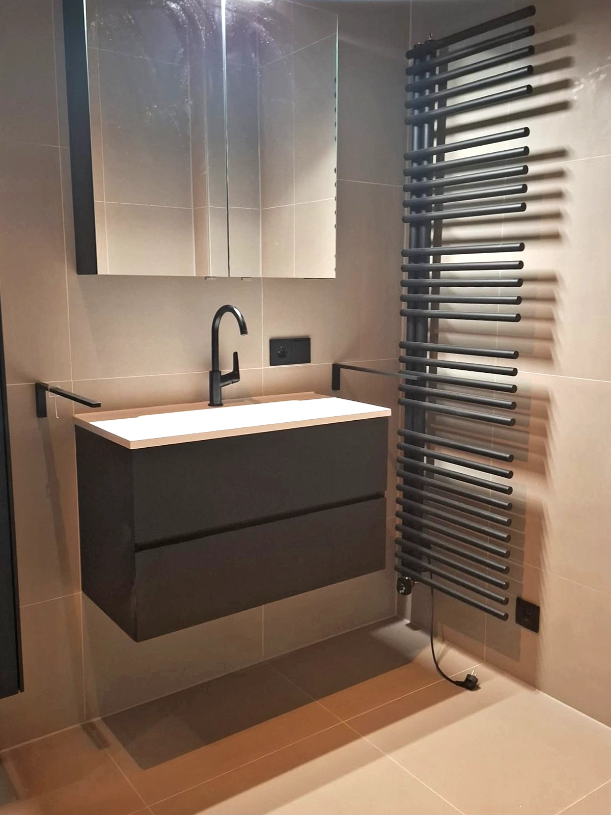 Moderne badkamer met donker badkamermeubel, zwarte kraan, spiegelkast, zwarte verwarming en beige tegels.