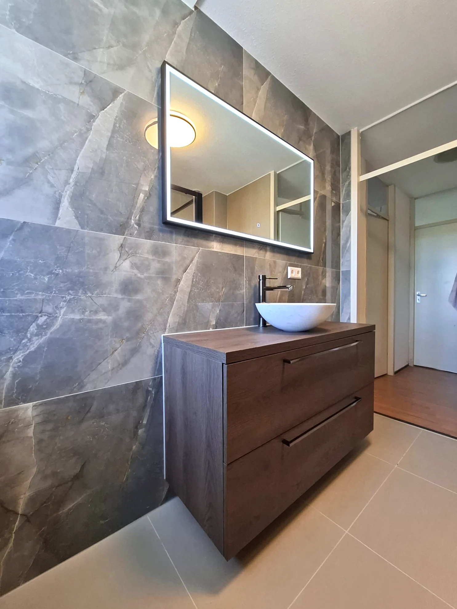 Modern badkamermeubel met houtlook afwerking, opbouwwastafel en verlichte spiegel tegen donkere marmerlook wandtegels.