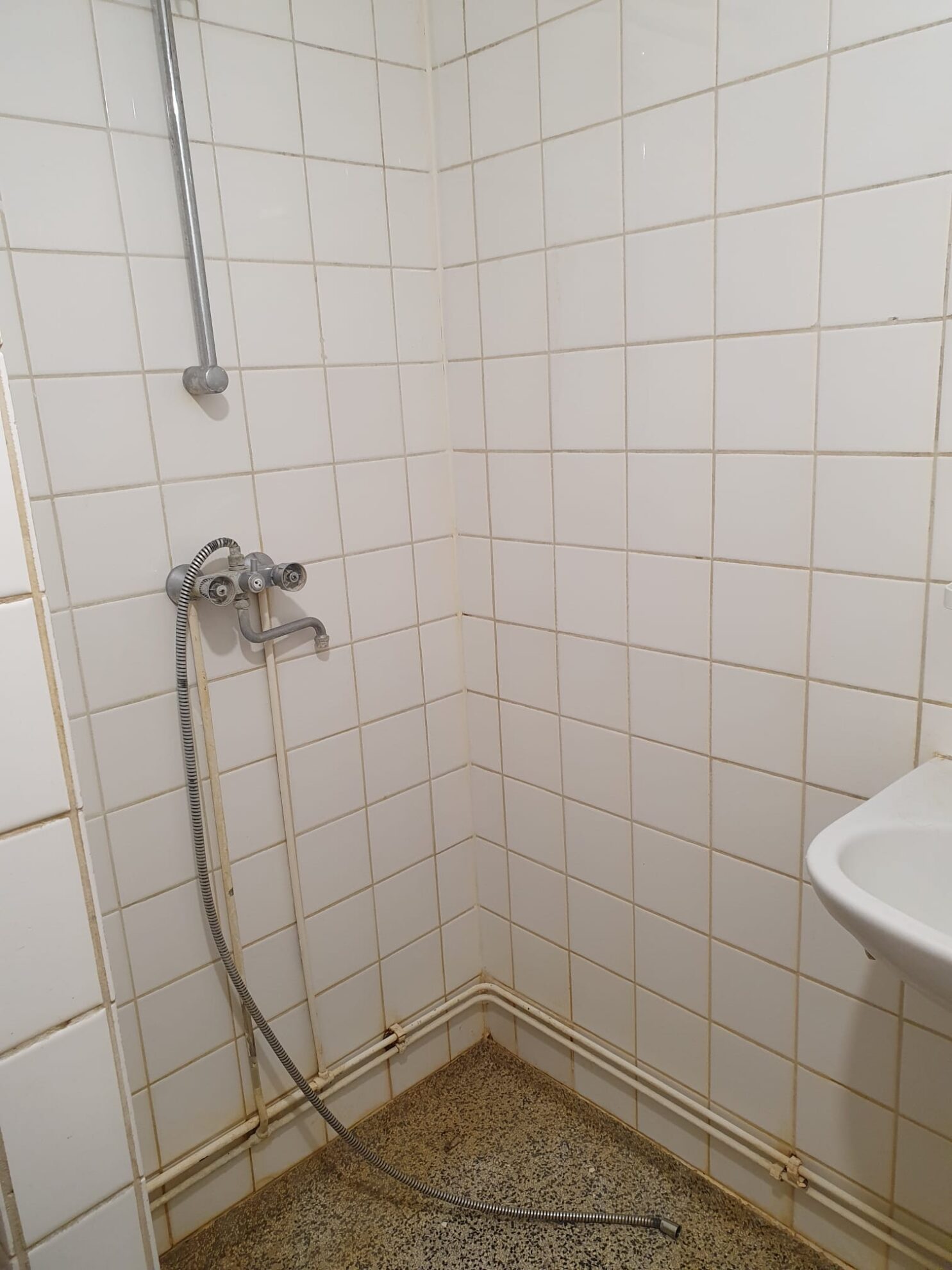 Badkamer vóór renovatie met douchehoek, witte wandtegels, opbouw douchekraan en zichtbaar leidingwerk.