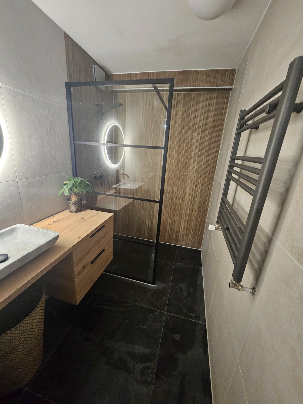 Moderne badkamer met inloopdouche, houtlook wandtegels, houten badkamermeubel, ronde verlichte spiegel en zwarte verwarming.