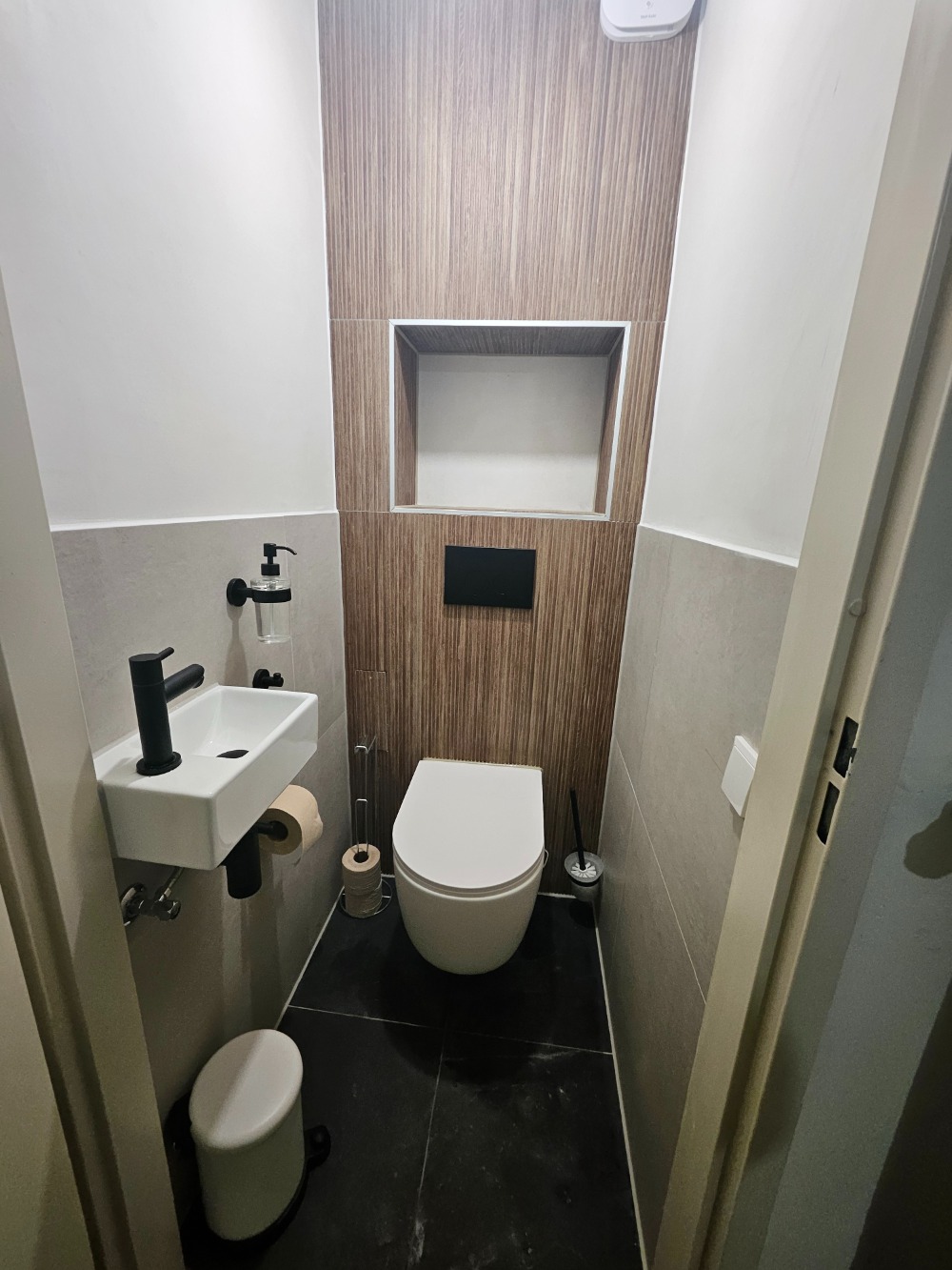 Moderne toiletruimte met wandtoilet, fontein, houtlook achterwand, wandnis en zwarte accessoires.