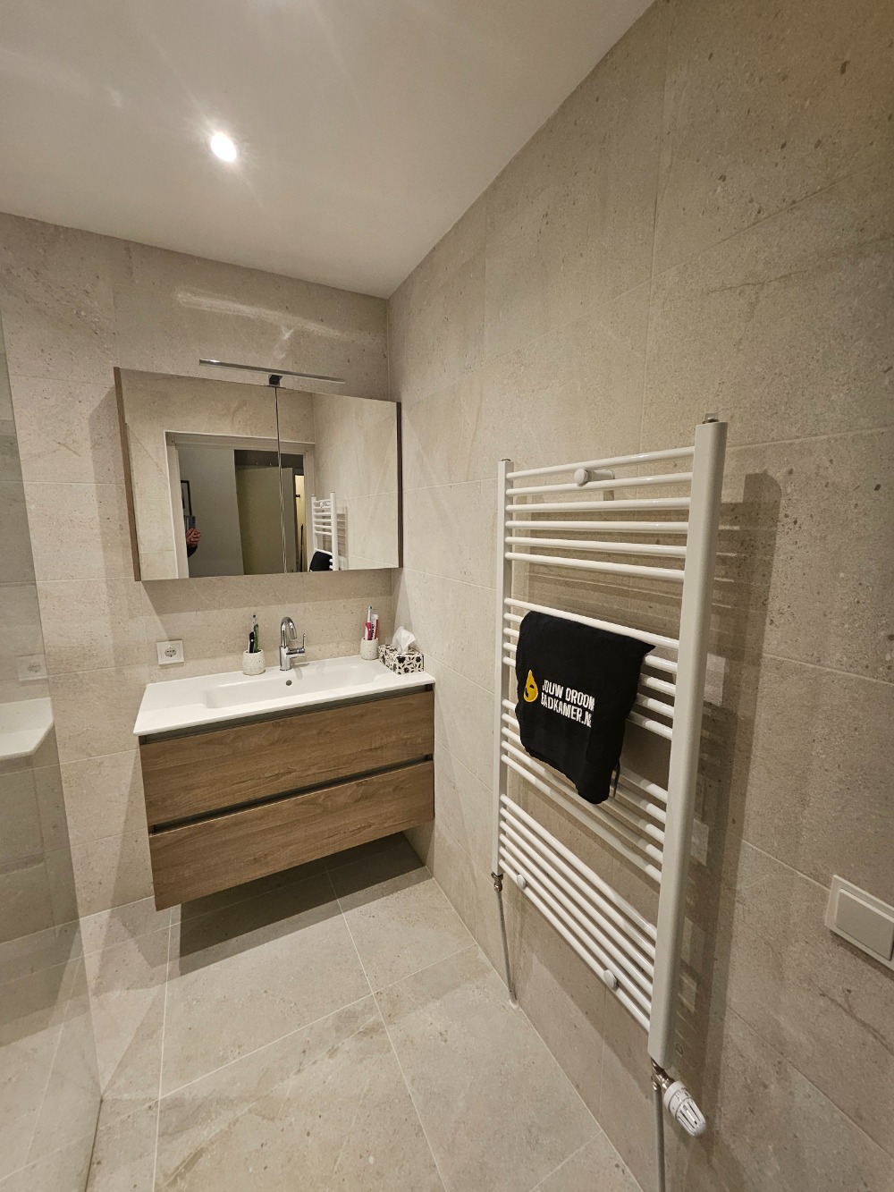 Moderne badkamer met houten badkamermeubel, spiegelkast, witte verwarming en beige wand- en vloertegels.