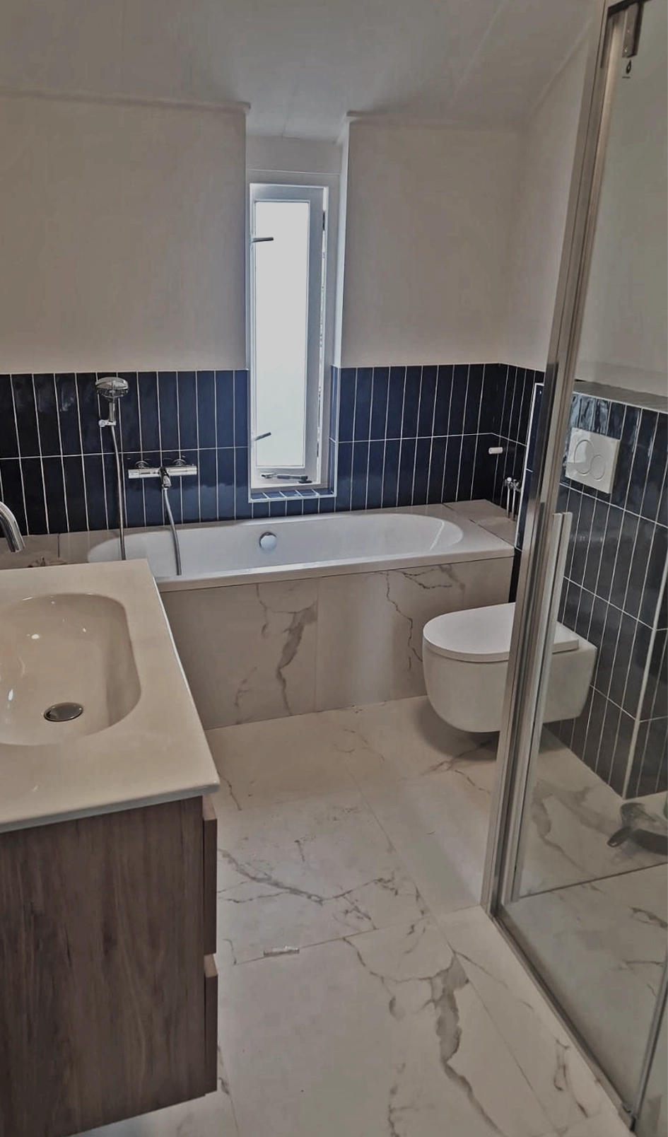 Moderne badkamer met ligbad, donkerblauwe wandtegels en marmerlook vloertegels.