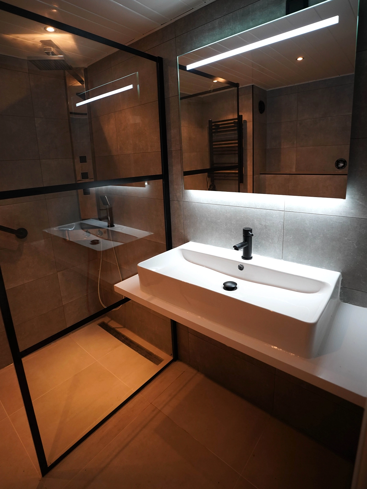Moderne badkamer met zwarte douchewand, verlichte spiegel en witte opbouwwastafel.