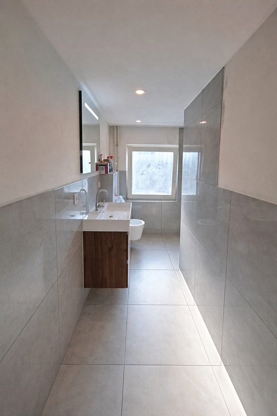 Smalle moderne badkamer met lichtgrijze tegels, houten wastafelmeubel en zwevend toilet.