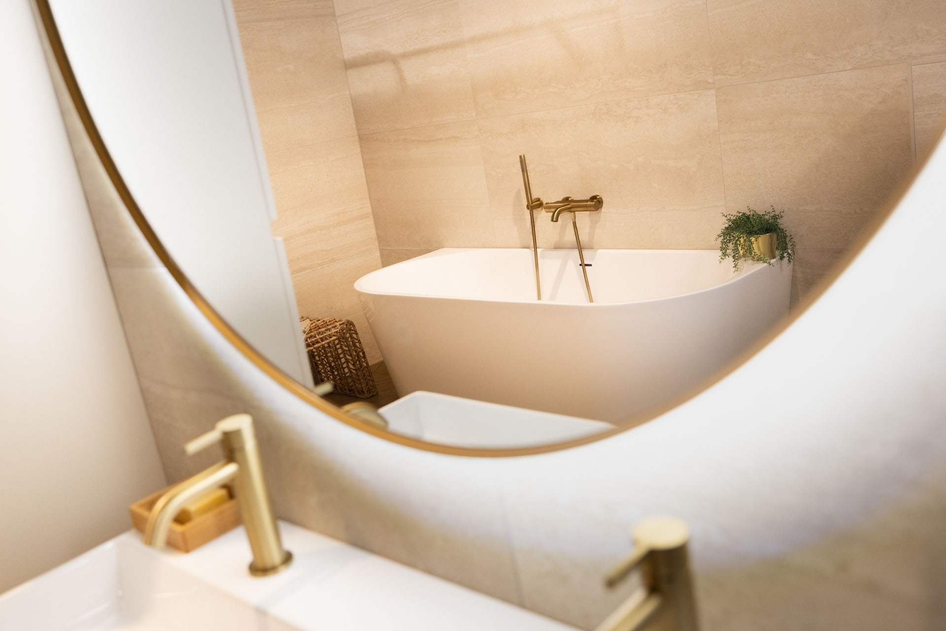 Vrijstaand ligbad met gouden kranen in een moderne badkamer met beige wandtegels.