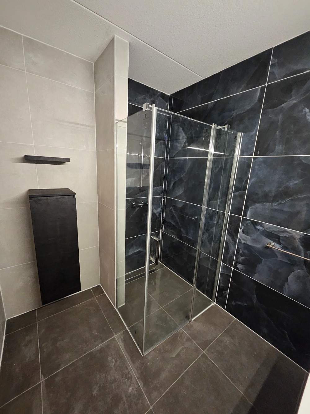 Moderne badkamer met glazen inloopdouche, donkere marmerlook wandtegels en strakke zwarte accessoires.