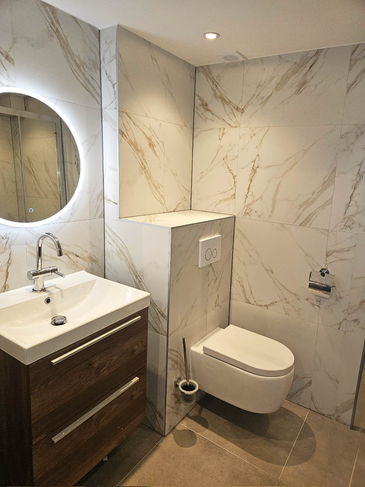 Moderne badkamer met marmerlook wandtegels, zwevend toilet en houten badkamermeubel met ronde spiegel.