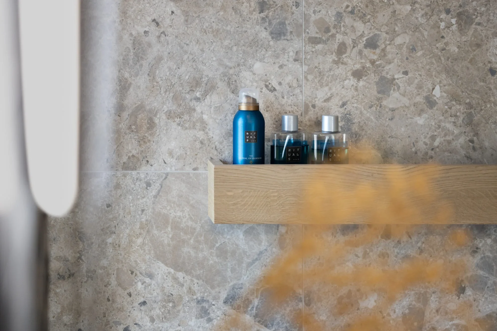 Houten doucheplank met verzorgingsproducten tegen grijze natuursteenlook wandtegels in een moderne badkamer.