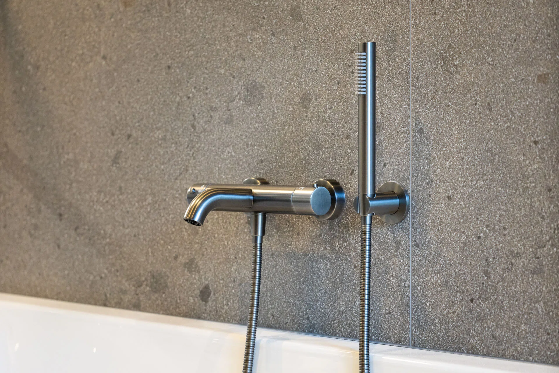 Geborsteld stalen badkraan met handdouche bij een wit bad tegen grijze wandtegels.