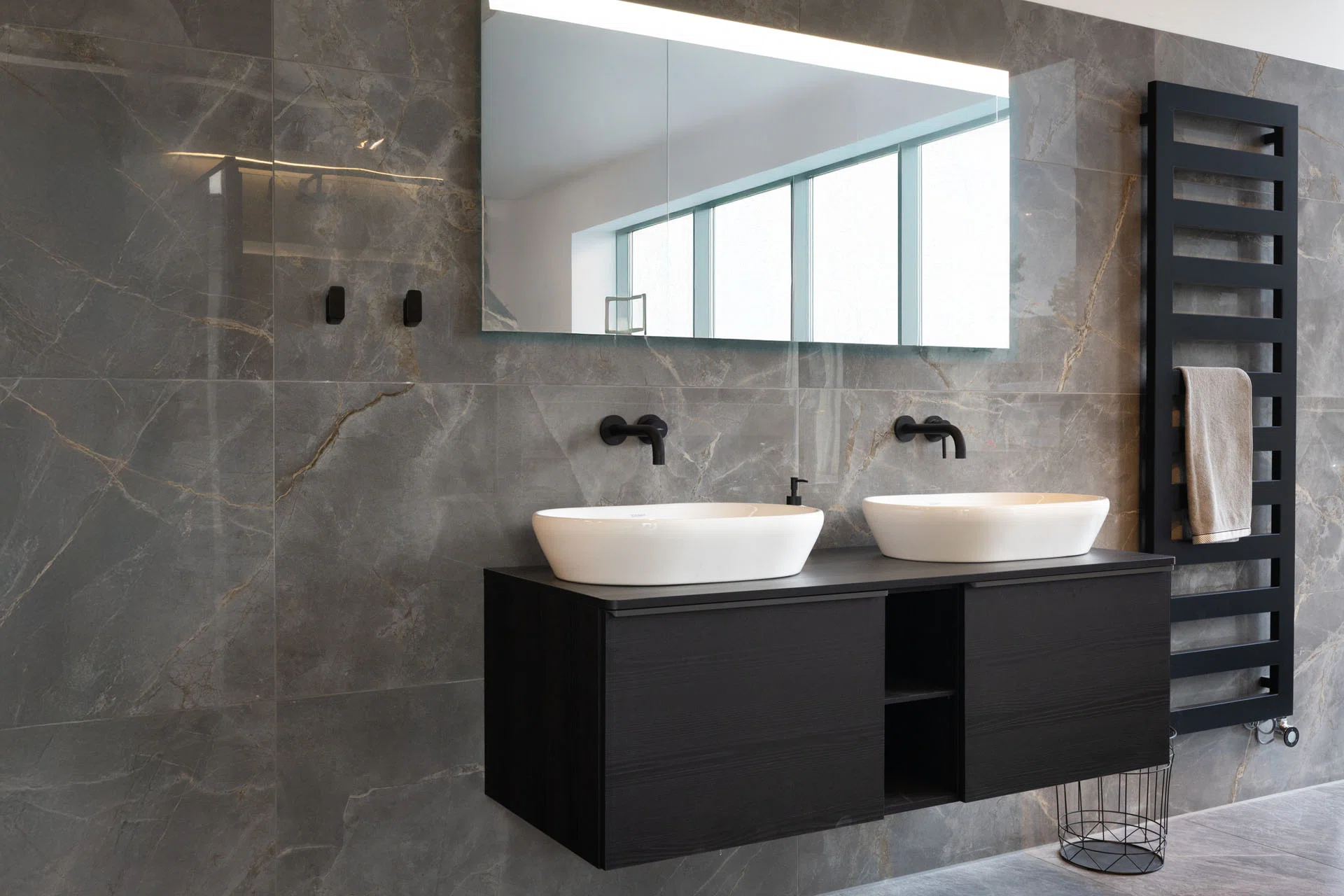 Luxe badkamer met dubbel badkamermeubel, witte waskommen, zwarte wandkranen en grijze marmerlook wandtegels.
