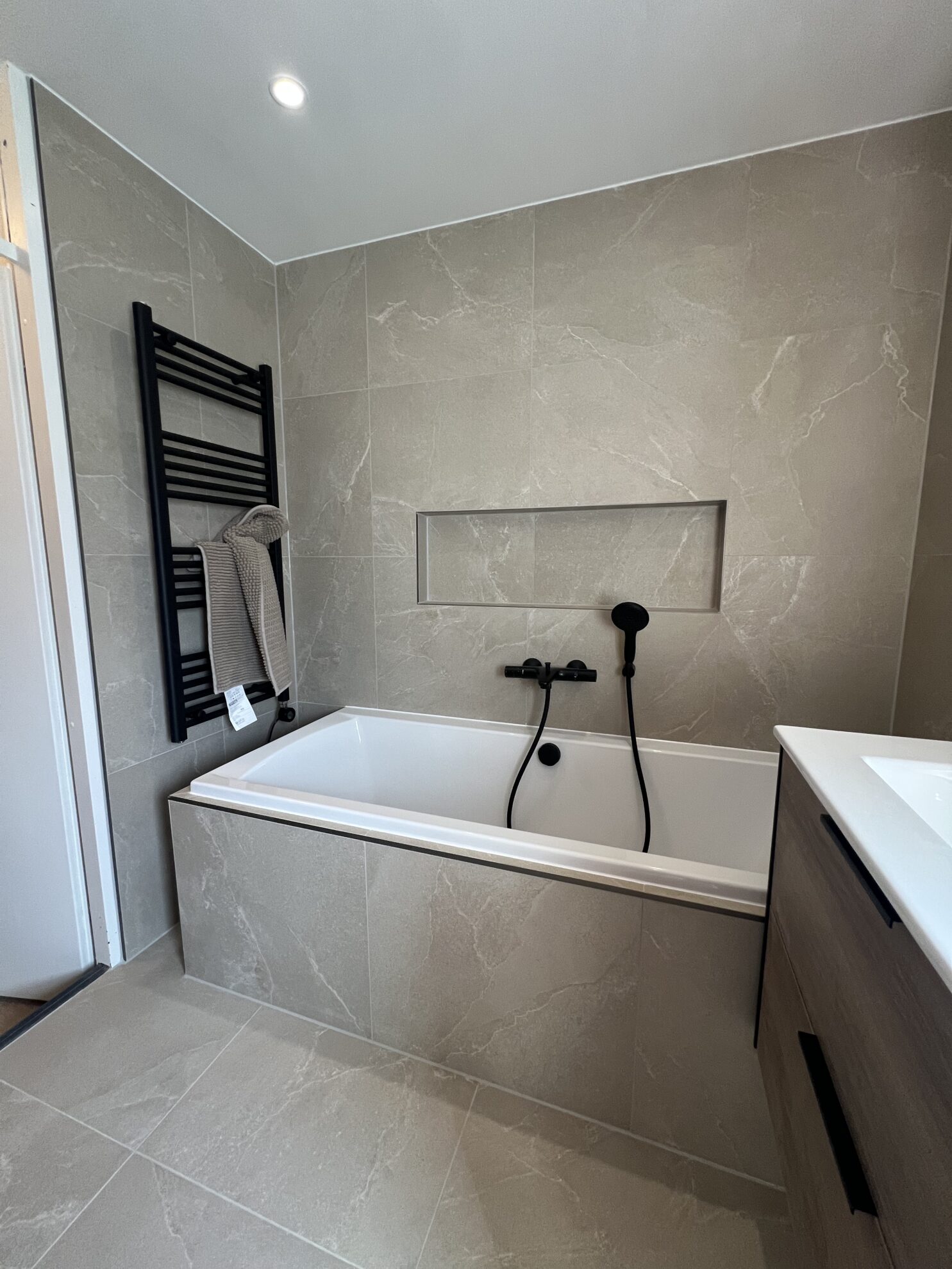 Modern ligbad in badkamer met grijze tegels, zwarte badkraan en handdoekradiator