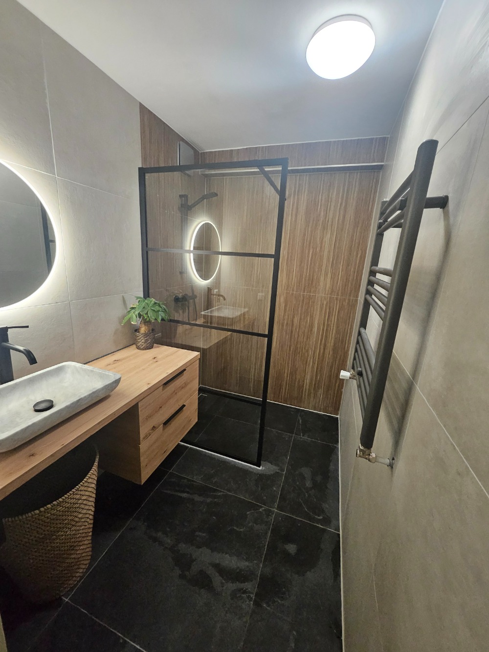Moderne badkamer met inloopdouche, houtlook wandtegels, houten badkamermeubel, ronde verlichte spiegel en zwarte verwarming.