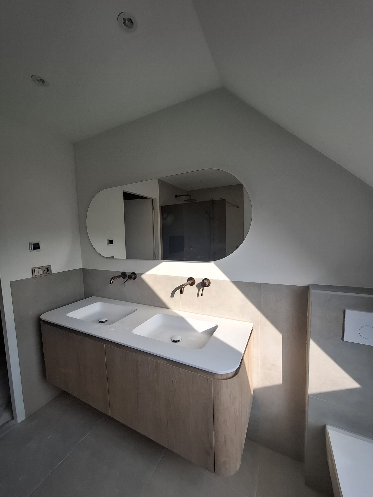 Moderne badkamer met dubbele wastafel, houten badkamermeubel en organische spiegel onder een schuin dak.