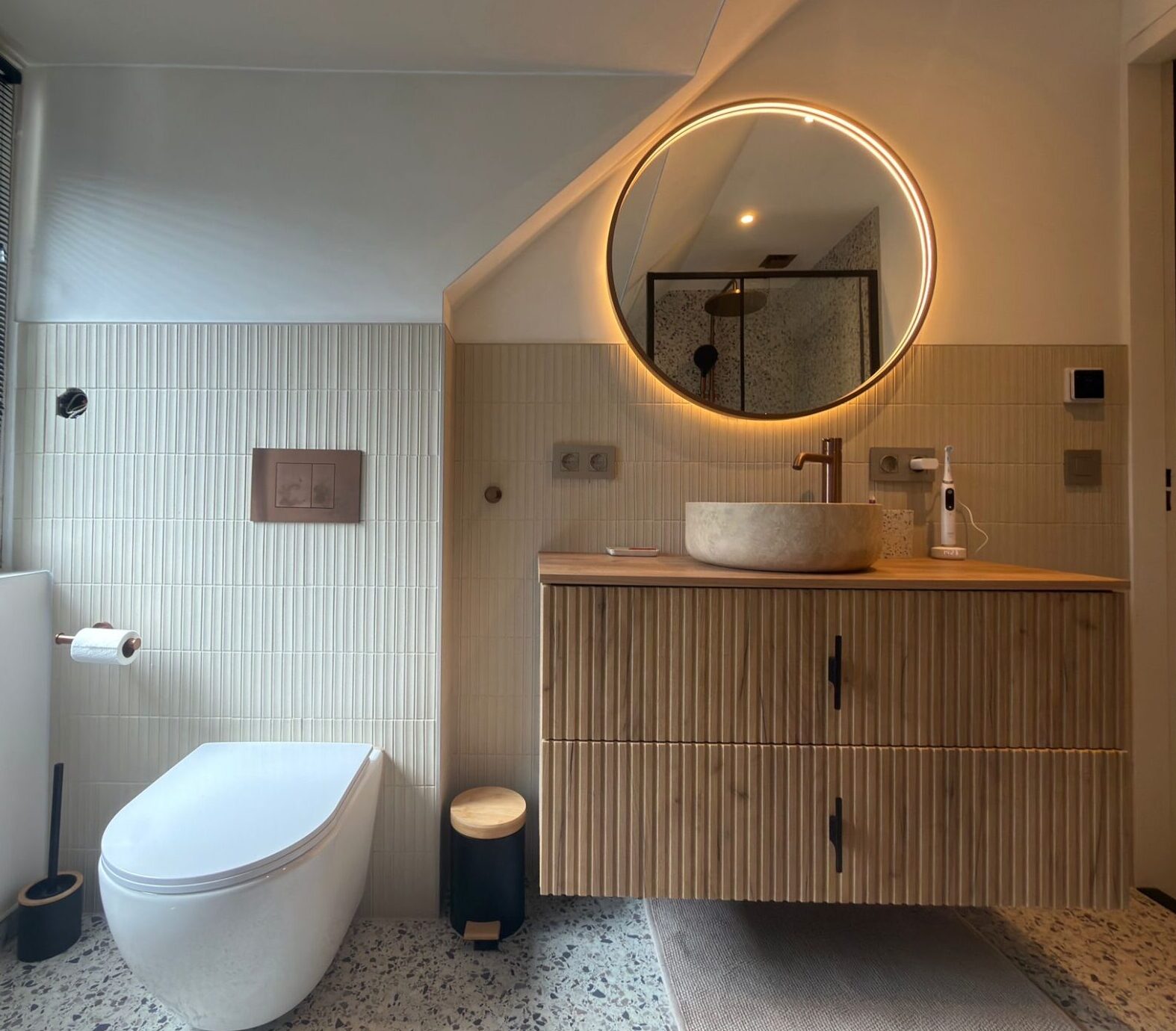 Modern toilet met houten fonteinmeubel, natuurstenen waskom, ronde verlichte spiegel en terrazzo vloer.