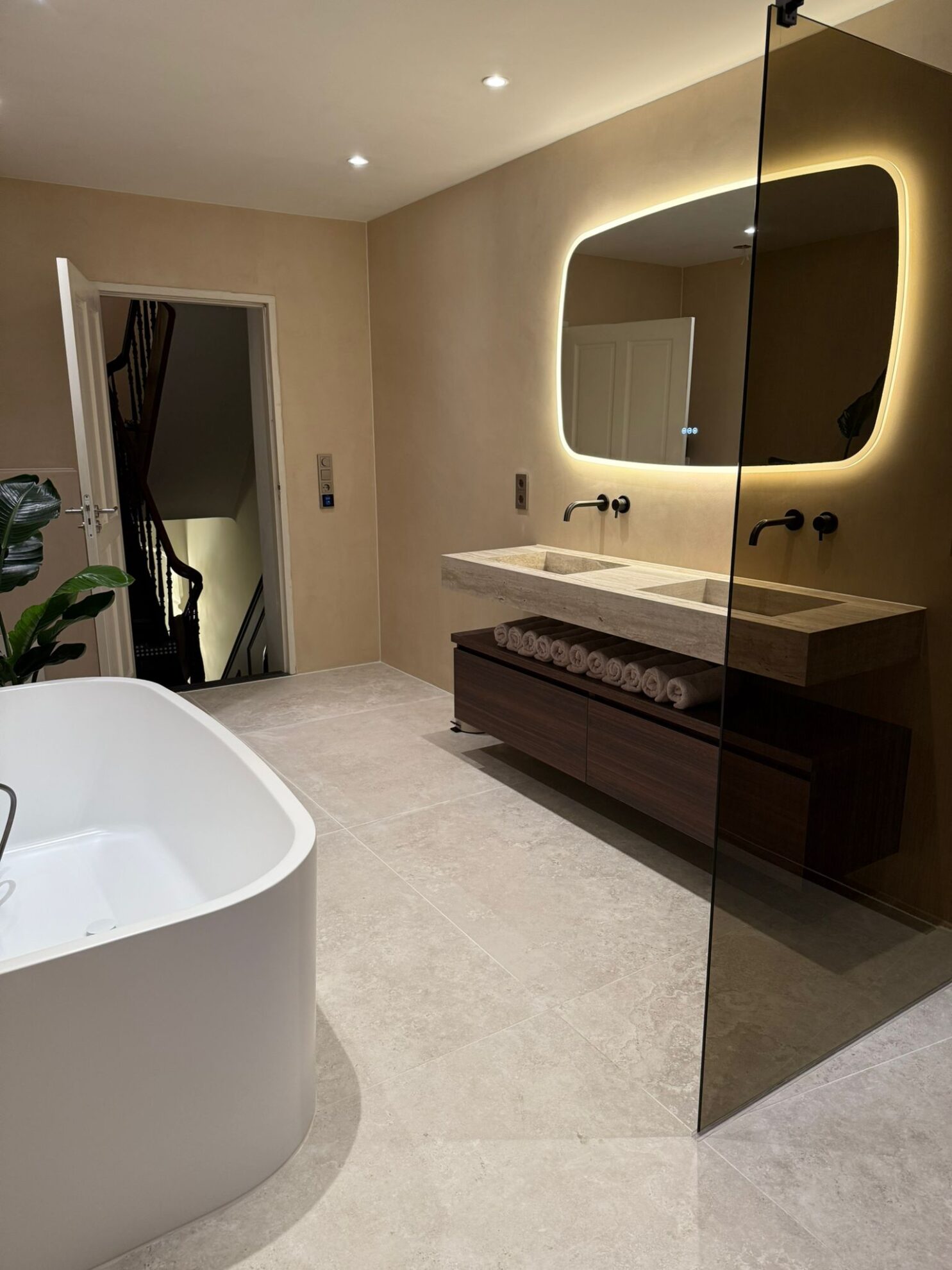 Luxe badkamer met vrijstaand ligbad, dubbele natuurstenen wastafel, verlichte spiegel en glazen inloopdouche.